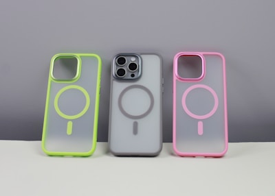 Three phone cases are displayed on a neutral background. The left case has a neon green border, the middle case contains a phone and has a gray border, while the right case features a pink border. Each case has a circular design in the center.