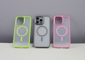 Three phone cases are displayed on a neutral background. The left case has a neon green border, the middle case contains a phone and has a gray border, while the right case features a pink border. Each case has a circular design in the center.