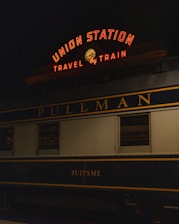 A vintage train car is parked at a station under a neon sign that reads 'Union Station Travel by Train'. The train car is labeled 'Pullman' and 'Suitsme', with a classic design typical of early 20th-century railcars.