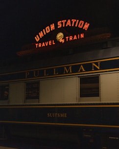 A vintage train car is parked at a station under a neon sign that reads 'Union Station Travel by Train'. The train car is labeled 'Pullman' and 'Suitsme', with a classic design typical of early 20th-century railcars.
