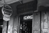A black-and-white photograph of a storefront with a sign reading 'Rev. Zombie's Voodoo Shop' offering readings and rituals. The entrance has weathered wooden doors and a slightly eerie appearance, with various flyers and signs posted nearby.