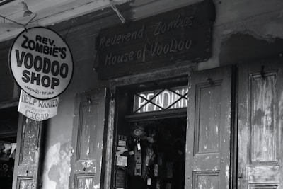 A black-and-white photograph of a storefront with a sign reading 'Rev. Zombie's Voodoo Shop' offering readings and rituals. The entrance has weathered wooden doors and a slightly eerie appearance, with various flyers and signs posted nearby.