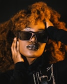 A person with voluminous curly hair, wearing stylish sunglasses and dark lipstick. The individual has their hand placed on their face and head, conveying a sense of confidence and poise. The attire includes dark clothing with metallic chain accessories, adding an element of edginess.