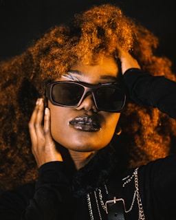 A person with voluminous curly hair, wearing stylish sunglasses and dark lipstick. The individual has their hand placed on their face and head, conveying a sense of confidence and poise. The attire includes dark clothing with metallic chain accessories, adding an element of edginess.