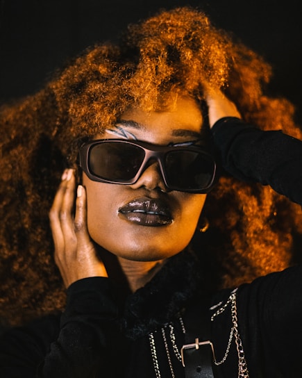 A person with voluminous curly hair, wearing stylish sunglasses and dark lipstick. The individual has their hand placed on their face and head, conveying a sense of confidence and poise. The attire includes dark clothing with metallic chain accessories, adding an element of edginess.