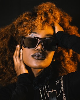 A person with voluminous curly hair, wearing stylish sunglasses and dark lipstick. The individual has their hand placed on their face and head, conveying a sense of confidence and poise. The attire includes dark clothing with metallic chain accessories, adding an element of edginess.