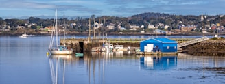 A peaceful waterfront scene with several small boats docked at a wooden pier. A bright blue boathouse stands prominently on the water's edge, reflecting in the calm, glassy surface of the water. In the background, a small town with colorful houses nestled among trees is visible, with a clear blue sky overhead.