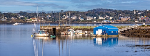 A peaceful waterfront scene with several small boats docked at a wooden pier. A bright blue boathouse stands prominently on the water's edge, reflecting in the calm, glassy surface of the water. In the background, a small town with colorful houses nestled among trees is visible, with a clear blue sky overhead.
