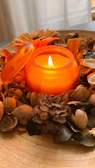 Burnt orange candle casting a warm glow on rough linen fabric with scattered pine needles.