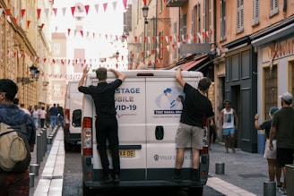 Two people are positioned on the back of a white van with lettering promoting COVID-19 nasal testing. They appear to be working or adjusting something on the vehicle. The street is decorated with red and white triangular flags strung overhead, and there are other people and vehicles visible in the background. The scene is set in a narrow European-style street with pastel-colored buildings on either side.