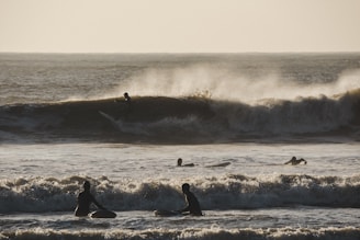 Surfers are riding waves on a rough sea, with the sun casting a golden light over the scene. The waves are large and powerful, creating dynamic splashes and spray as they break. Silhouetted surfers in the foreground are sitting on their boards, waiting for a wave, while one surfer is visibly riding a wave in the background.