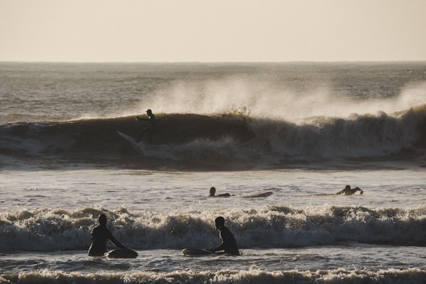 Surfers are riding waves on a rough sea, with the sun casting a golden light over the scene. The waves are large and powerful, creating dynamic splashes and spray as they break. Silhouetted surfers in the foreground are sitting on their boards, waiting for a wave, while one surfer is visibly riding a wave in the background.