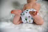 A baby lies on a soft, fluffy white rug, wearing a cloth diaper with a cactus pattern. The focus is on the baby's legs and feet, showing a sense of comfort and innocence.