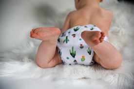 A baby lies on a soft, fluffy white rug, wearing a cloth diaper with a cactus pattern. The focus is on the baby's legs and feet, showing a sense of comfort and innocence.
