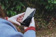 A person holds a smartphone displaying a loading screen for Adobe Photoshop with a progress circle. The background consists of blurred green foliage, suggesting an outdoor setting. The individual wears a long sleeve garment, likely a sweater, in a muted blue shade with a visible red cuff.