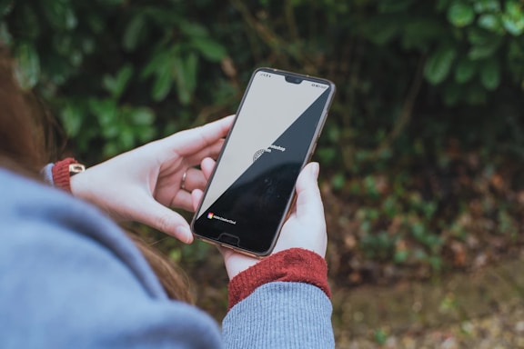 A person holds a smartphone displaying a loading screen for Adobe Photoshop with a progress circle. The background consists of blurred green foliage, suggesting an outdoor setting. The individual wears a long sleeve garment, likely a sweater, in a muted blue shade with a visible red cuff.