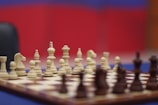 A chessboard set up for a game with white pieces in focus and a blurred background featuring red and blue colors. The chess pieces are arranged for an opening move.