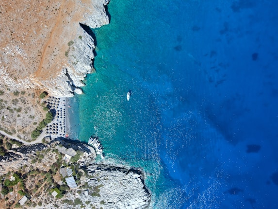 Aerial view of a coastal area with clear turquoise waters meeting rocky cliffs. A sandy beach with umbrellas is situated near the shoreline. The blue of the sea contrasts with the browns and greens of the land. Several boats are anchored close to the shore.