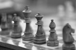 A close-up view of a chessboard featuring wooden chess pieces. The focus is on the king piece, surrounded by other pieces such as a bishop and a knight. The image is in black and white, giving it a classic, timeless feel.