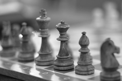 A close-up view of a chessboard featuring wooden chess pieces. The focus is on the king piece, surrounded by other pieces such as a bishop and a knight. The image is in black and white, giving it a classic, timeless feel.