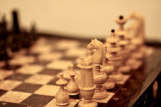 A close-up view of a chessboard with intricately carved wooden chess pieces. The focus is on the knight piece with its detailed mane, surrounded by pawns, rooks, and bishops. The chessboard squares are inlaid with light and dark wood.