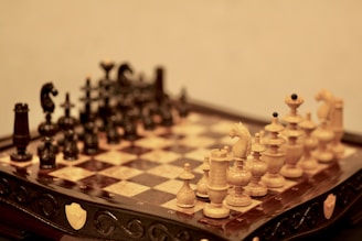 A detailed view of a chessboard with intricately carved chess pieces in a classical design. The board is made of dark and light squares with ornamental wooden craftsmanship visible along the edges. The pieces are arranged in their starting positions, with one set in a dark color and the other in a lighter shade, creating a contrasting visual.