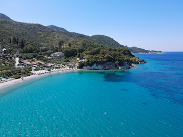 A beautiful coastal landscape featuring a serene beach with bright turquoise waters. The beach is lined with umbrellas and sun loungers, hinting at a popular relaxation spot for visitors. Surrounding hills are lush and green, creating a picturesque backdrop. The sky is clear, adding to the tranquil and inviting atmosphere.