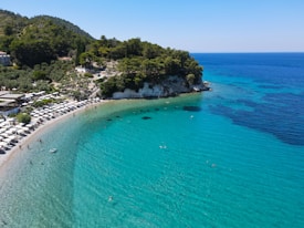 A picturesque beach with clear turquoise waters gently lapping against the shore. Visitors enjoy sunbathing on neatly organized loungers under umbrellas. Lush green hills provide a scenic backdrop to the idyllic coastal scene, with a few people swimming and playing in the water.