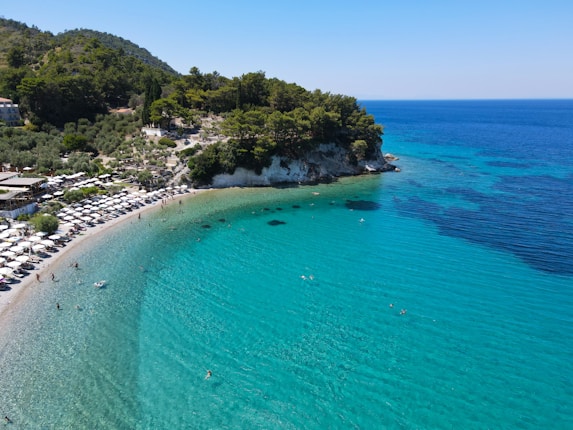 A picturesque beach with clear turquoise waters gently lapping against the shore. Visitors enjoy sunbathing on neatly organized loungers under umbrellas. Lush green hills provide a scenic backdrop to the idyllic coastal scene, with a few people swimming and playing in the water.