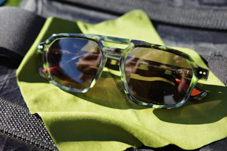 A pair of sunglasses with a tortoiseshell frame rests on a bright green cleaning cloth. The lenses reflect some outdoor scenery and the sunglasses are placed on a textured dark surface.