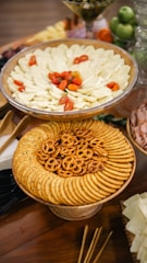 A table features an arrangement of various appetizers, including a large platter with sliced cheese and cherry tomatoes, another plate with round crackers and pretzels, and a glass filled with green apples and other fruits in the background.