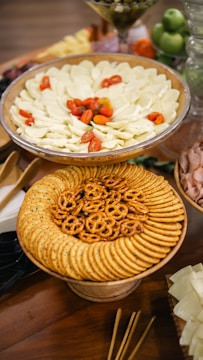 A table features an arrangement of various appetizers, including a large platter with sliced cheese and cherry tomatoes, another plate with round crackers and pretzels, and a glass filled with green apples and other fruits in the background.