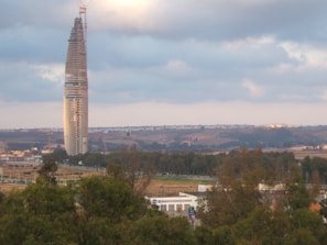 A tall, slender skyscraper under construction stands prominently against a backdrop of hills and cloudy skies. The scene includes surrounding greenery and urban infrastructure at the ground level.