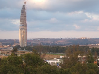 A tall, slender skyscraper under construction stands prominently against a backdrop of hills and cloudy skies. The scene includes surrounding greenery and urban infrastructure at the ground level.