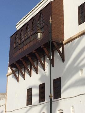 A traditional architectural design featuring a white building with a wooden overhanging balcony supported by wooden beams. The building has arched windows and a streetlamp is positioned in front of it. The bright sky is visible in the background.