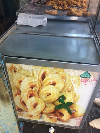 An outdoor food stall counter with metal surfaces and a large image of donuts displayed on the front. Several freshly fried dough items, including what appear to be churros and other pastries, are placed on trays on top of the counter. A crumpled paper bag is nearby, and some debris is scattered across the surface.
