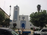 A white building with blue accents, featuring a large cross and clock on its facade. It is surrounded by trees and several people are visible near the entrance. Vehicles are parked in the foreground.