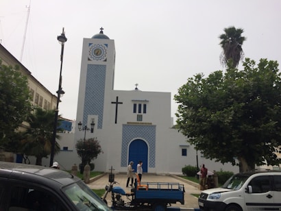 A white building with blue accents, featuring a large cross and clock on its facade. It is surrounded by trees and several people are visible near the entrance. Vehicles are parked in the foreground.