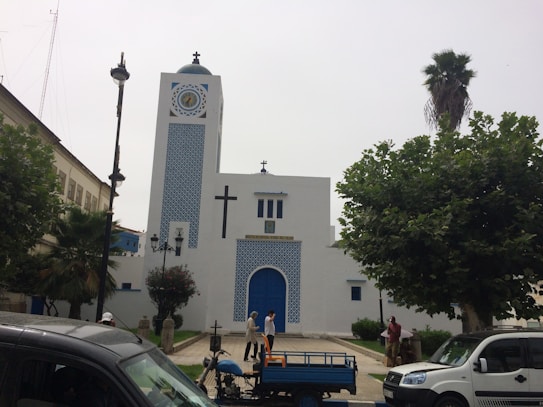 A white building with blue accents, featuring a large cross and clock on its facade. It is surrounded by trees and several people are visible near the entrance. Vehicles are parked in the foreground.