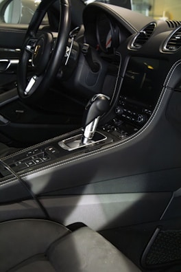 The interior of a luxury car featuring a sleek black steering wheel, a gearshift, and a modern dashboard with a digital display. The seats and console are upholstered in dark leather, with detailed stitching visible.