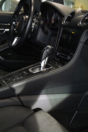 The interior of a luxury car featuring a sleek black steering wheel, a gearshift, and a modern dashboard with a digital display. The seats and console are upholstered in dark leather, with detailed stitching visible.