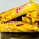 Three snack bars packaged in bright yellow wrappers are arranged, partially overlapping each other. The text on the wrappers indicates the brand and the word 'Original'. There are images of cereal pieces on the packaging. The bars seem to be crunchy cereal snacks likely from a known brand.