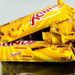 Three snack bars packaged in bright yellow wrappers are arranged, partially overlapping each other. The text on the wrappers indicates the brand and the word 'Original'. There are images of cereal pieces on the packaging. The bars seem to be crunchy cereal snacks likely from a known brand.
