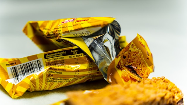 A close-up view of two crumpled, yellow snack wrappers with visible nutritional information in multiple languages. Pieces of a cereal bar are visible in the foreground.