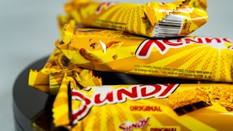 Several yellow-wrapped chocolate bars with a distinctive red logo and images of waffle texture on the packaging are stacked together on a reflective black surface.