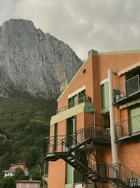 A multi-story building with orange and green accents stands prominently against the backdrop of a towering, rocky mountain. The structure features balconies with black railing and external staircases. Lush greenery surrounds the area, with parts of the mountain covered in trees. Other residential buildings are visible in the distance.