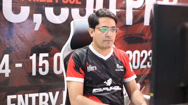 A man with glasses is sitting in a gaming chair, focused on a monitor in front of him. He is wearing a black and red esports jersey with various logos. The background features a large banner with text and numbers in bold fonts.