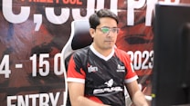 A man with glasses is sitting in a gaming chair, focused on a monitor in front of him. He is wearing a black and red esports jersey with various logos. The background features a large banner with text and numbers in bold fonts.