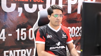 A man with glasses is sitting in a gaming chair, focused on a monitor in front of him. He is wearing a black and red esports jersey with various logos. The background features a large banner with text and numbers in bold fonts.
