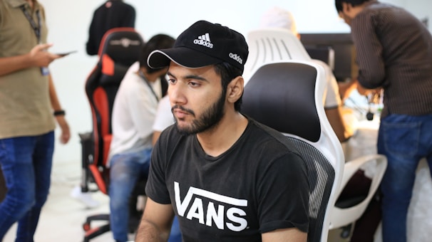 A group of young people are working in a modern office setting with computers. The focus is on a man wearing a black Adidas cap and a black Vans T-shirt, seated on a black and white chair. Others in the background also work on computers, with some standing and one holding a smartphone.